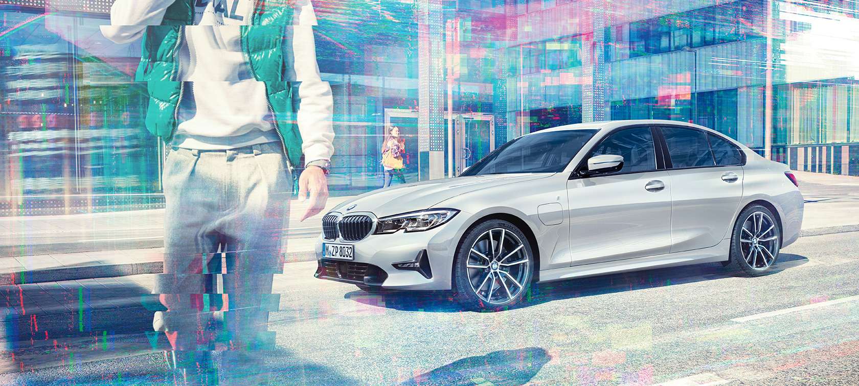 BMW 3 Series Sedan, side shot BMW 330e. G20 Three-quarter side shot of the standing BMW 330e Sedan in front of an urban glass building with walking passengers. G20