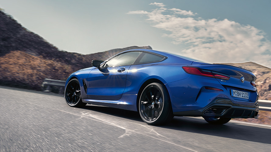 The BMW 8 Series Coupé G15 LCI Facelift three-quarter rear view driving The BMW 8 Series Coupé M Portimao Blue metallic G15 LCI Facelift three-quarter rear view driving in front of hills