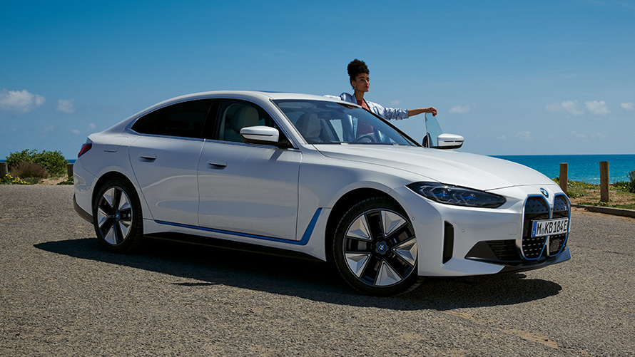 Woman standing in front of the open door of a BMW i4 in Mineral White G26 three-quarter side view  Woman standing in front of the open door of a BMW i4 in Mineral White G26 three-quarter side view