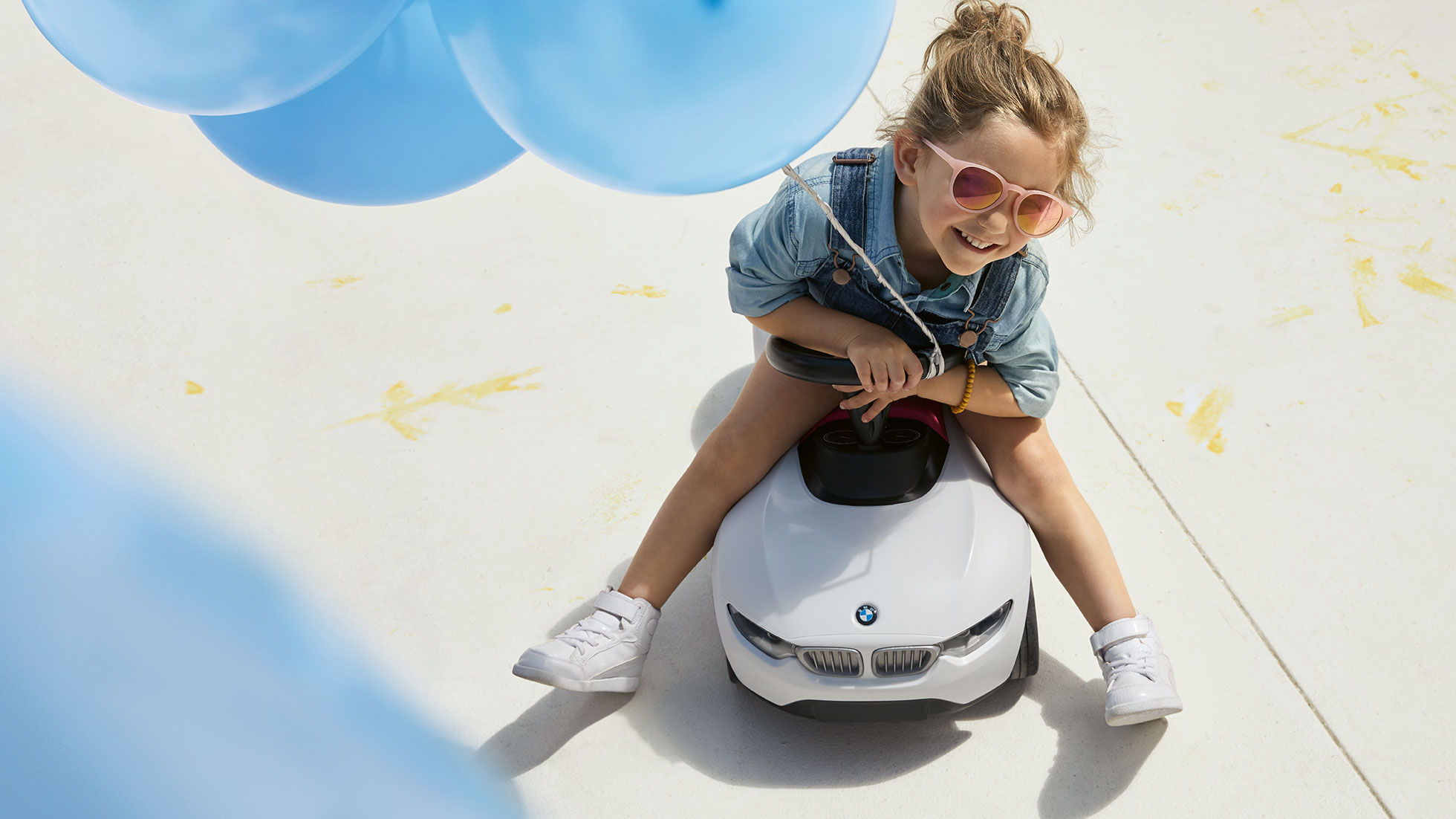 BMW Kids Collection. A girl with pink sunglasses and a blue balloon sits happily on a BMW Baby Racer.
